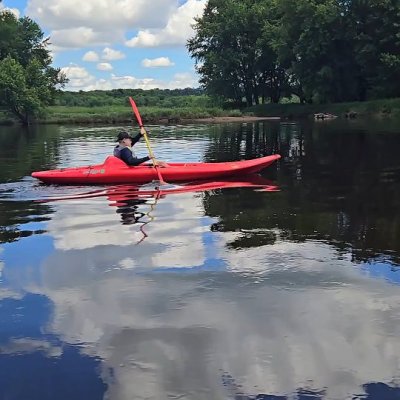 Person kayaking on calm water with trees and clouds reflected.