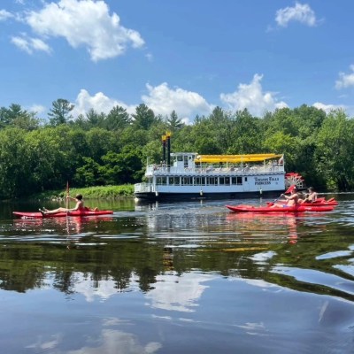 People kayaking near a white tour boat on a calm river with trees and clouds in the background.