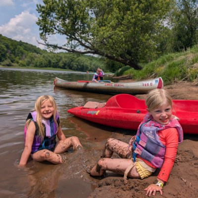 Two children in life jackets play by a lake near a canoe and kayak, surrounded by trees.
